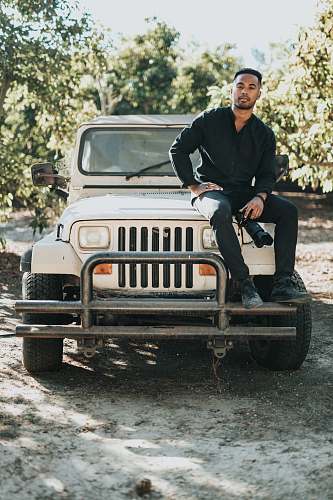 human man sitting on white Jeep Wrangler SUV near trees during daytime people
