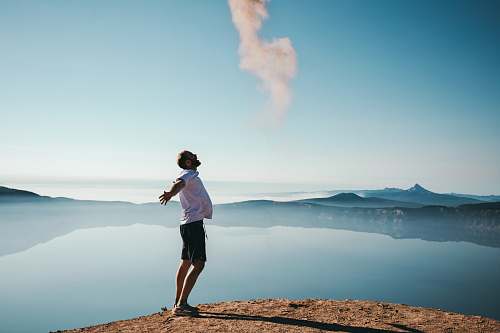 human man standing on sand while spreading arms beside calm body of water people