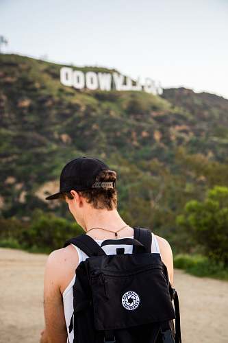 people man wearing white tank top and black snapback cap carrying black backpack in distance hollywood signboard at daytime human