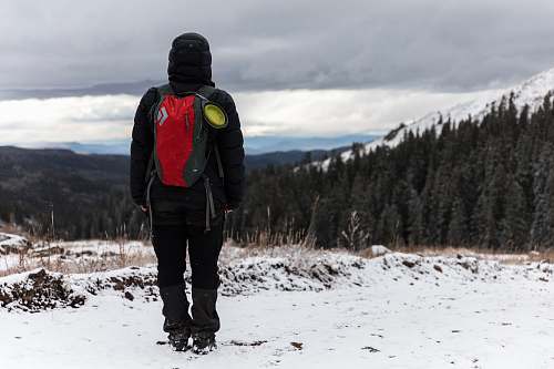 human person in black hoodie wearing red hydration pack and standing on top of hill covered of snow during cloudy sky people