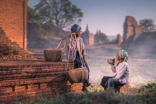 people sitting person smiling beside man sitting on brown concrete pavement while carrying baskets on his left shoulder human