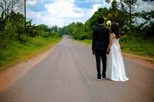suit wedding couple standing on winding road between trees under cumulus cloud road