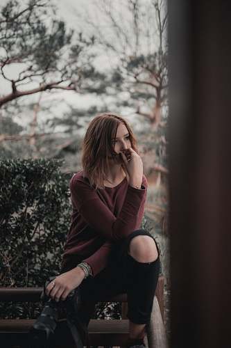 human wide-aperture photography of woman sitting on beige wooden frame near green shrub under gray clouded sky during daytime people