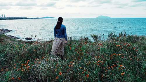 people woman in blue blouse and gray skirt standing on red flower field beside body of water during daytime human