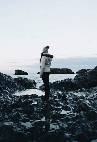 people woman in white parka jacket and black pants outfit standing on rocks near body of water under white sky at daytime human