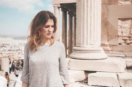 human woman wearing gray long-sleeved blouse standing beside white concrete column during daytime people