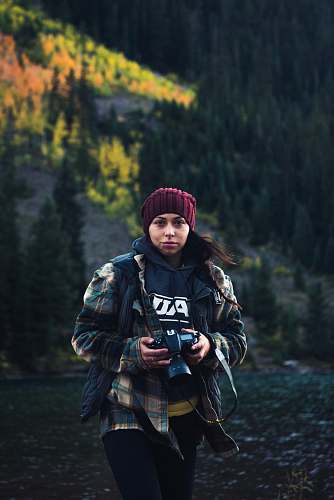human women wearing jacket and red knit cap holding DSLR camera walking near trees people