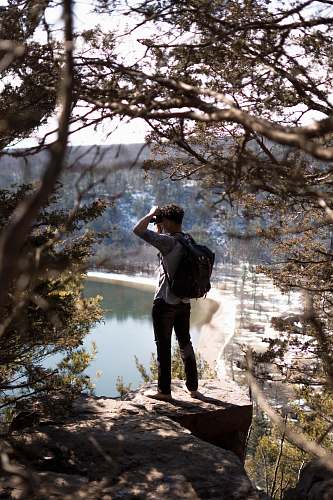 people man in gray dress shirt and black bag standing on rock formation while taking photo in front lake person