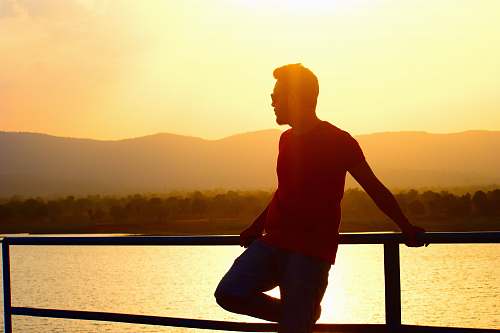 person man in red t-shirt and black pants leaning on railing in beach at sunset outdoors