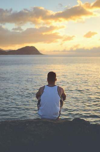person man sitting on rock near body of water during daytime apparel