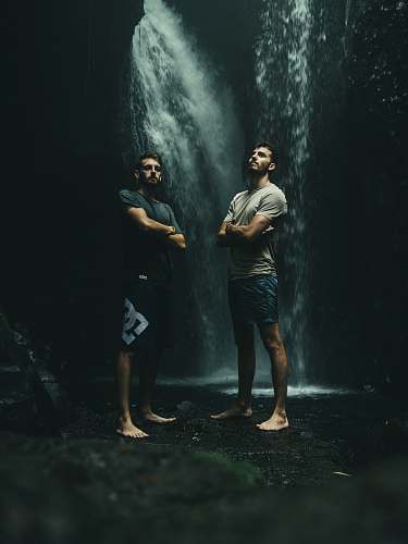 person two men standing at bottom of waterfall water