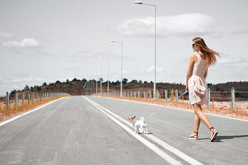 people woman in white sleeveless dress holding harness of puppy walking on asphalt road during daytime woman