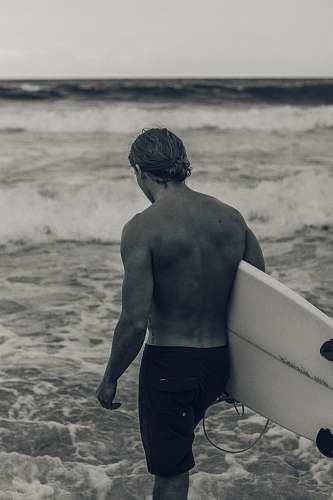 outdoors grayscale photography of man carrying a surfboard by the beach sea