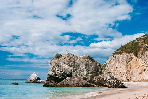 beach man standing on top on rock formation surrounded by body of water during daytime coast