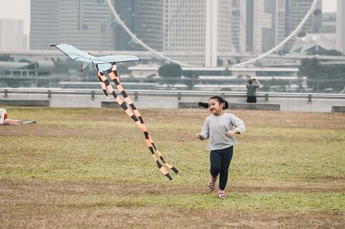 human girl swearing gray crew-neck long-sleeved shirt running in green field near orange and white plane kite viewing high-rise buildings sport