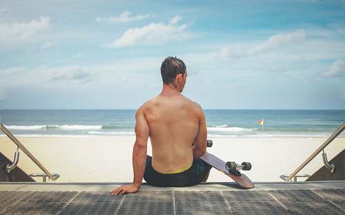 beach man sitting on gray surface holding skateboard ocean
