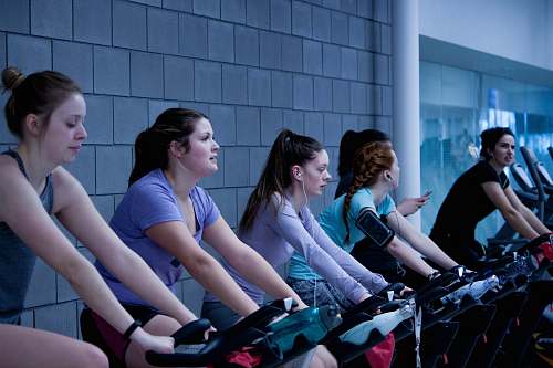 human women taking exercise on black stationary bikes in front of gray concrete wall people